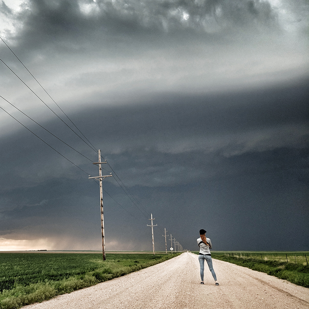 A Meteorologist standing under a thunderstorm on a gravel road next to telephone poles.