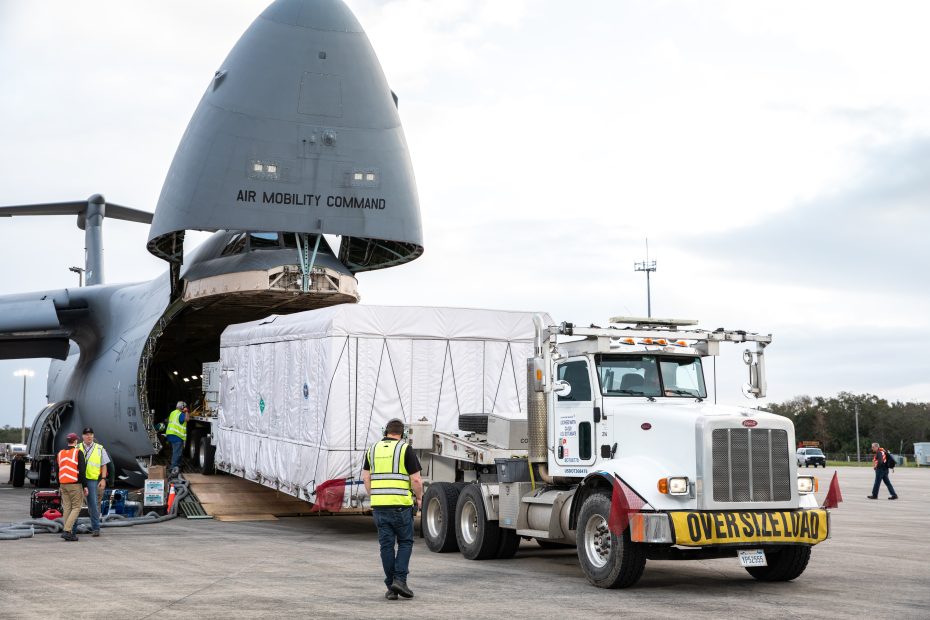 A picture of a large truck transferring a large weather satellite from an aircraft.
