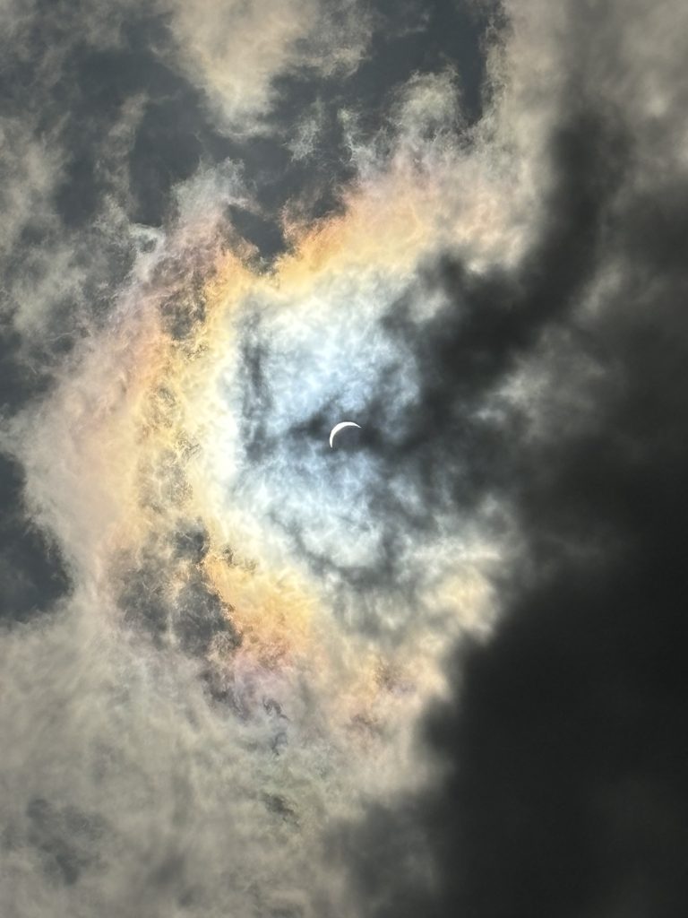 Image depicting the solar eclipse at approximately 80% coverage in Dallas, Texas, with clouds enveloping the sun, creating a mesmerizing rainbow hue around its edges. The partial eclipse is visible through breaks in the clouds, casting a soft glow over the landscape. The unique atmospheric conditions add a surreal and ethereal quality to the celestial event. Captured by Meteorologist Shannon Holland.