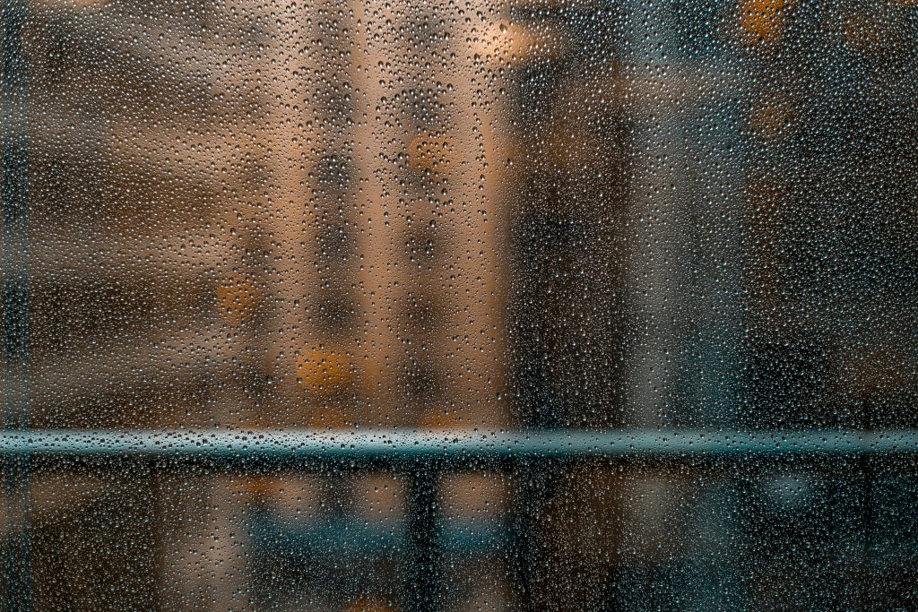 A photograph of rain droplets rolling down a glass window with high rise buildings in the background.