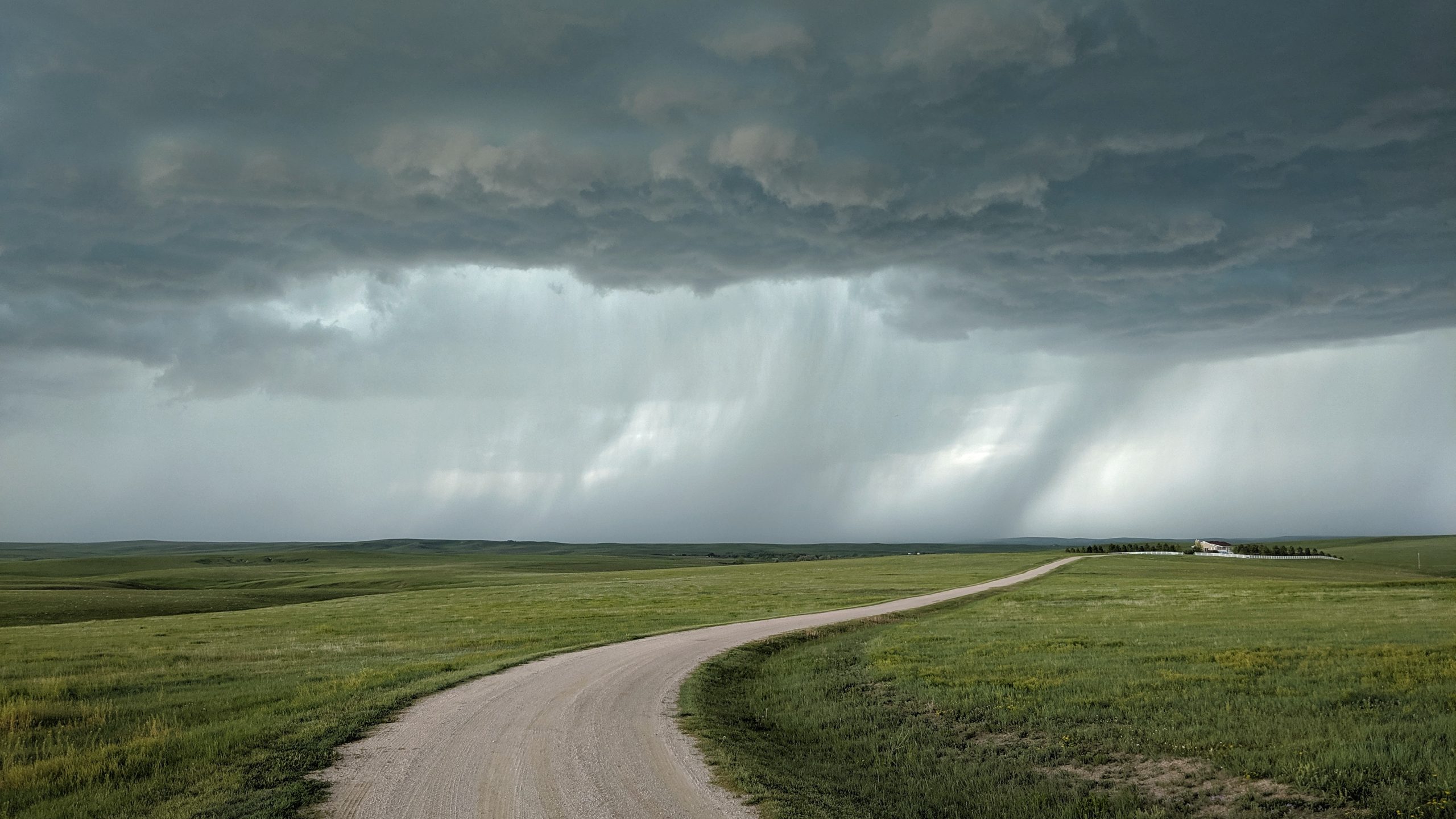 A photograph of a springtime thunderstorm and rain curtain over a green field with a house at the end of gravel road.
