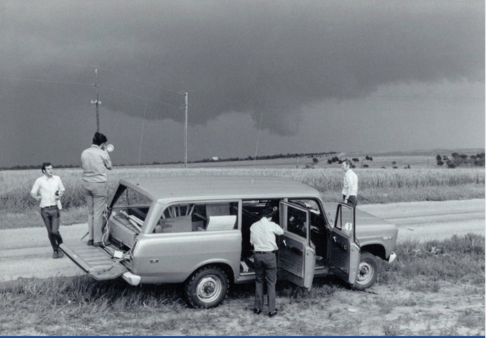 A black and white photograph of a NOAA field tornado research team standing around a vehicle on a dirt road photographing the development of a tornado.