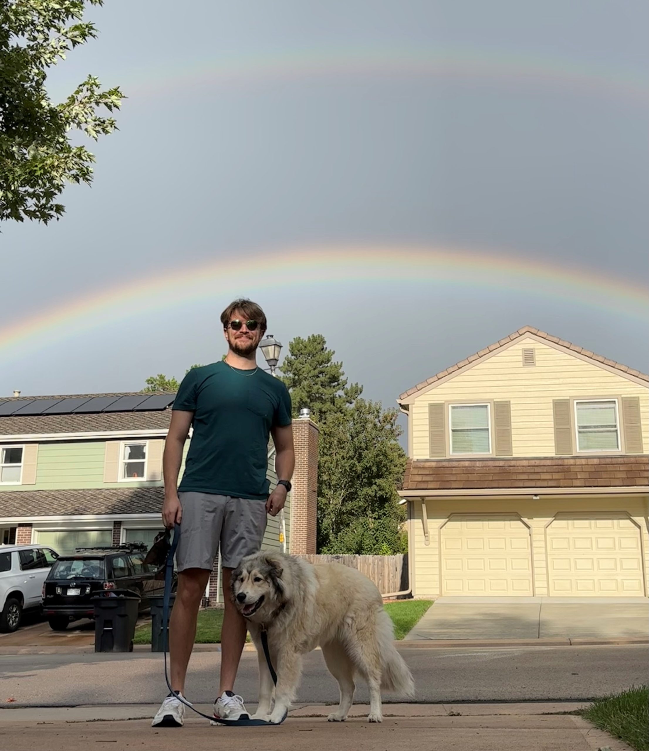 A man and a dog standing in a suburban neighborhood with a rainbow in the background.